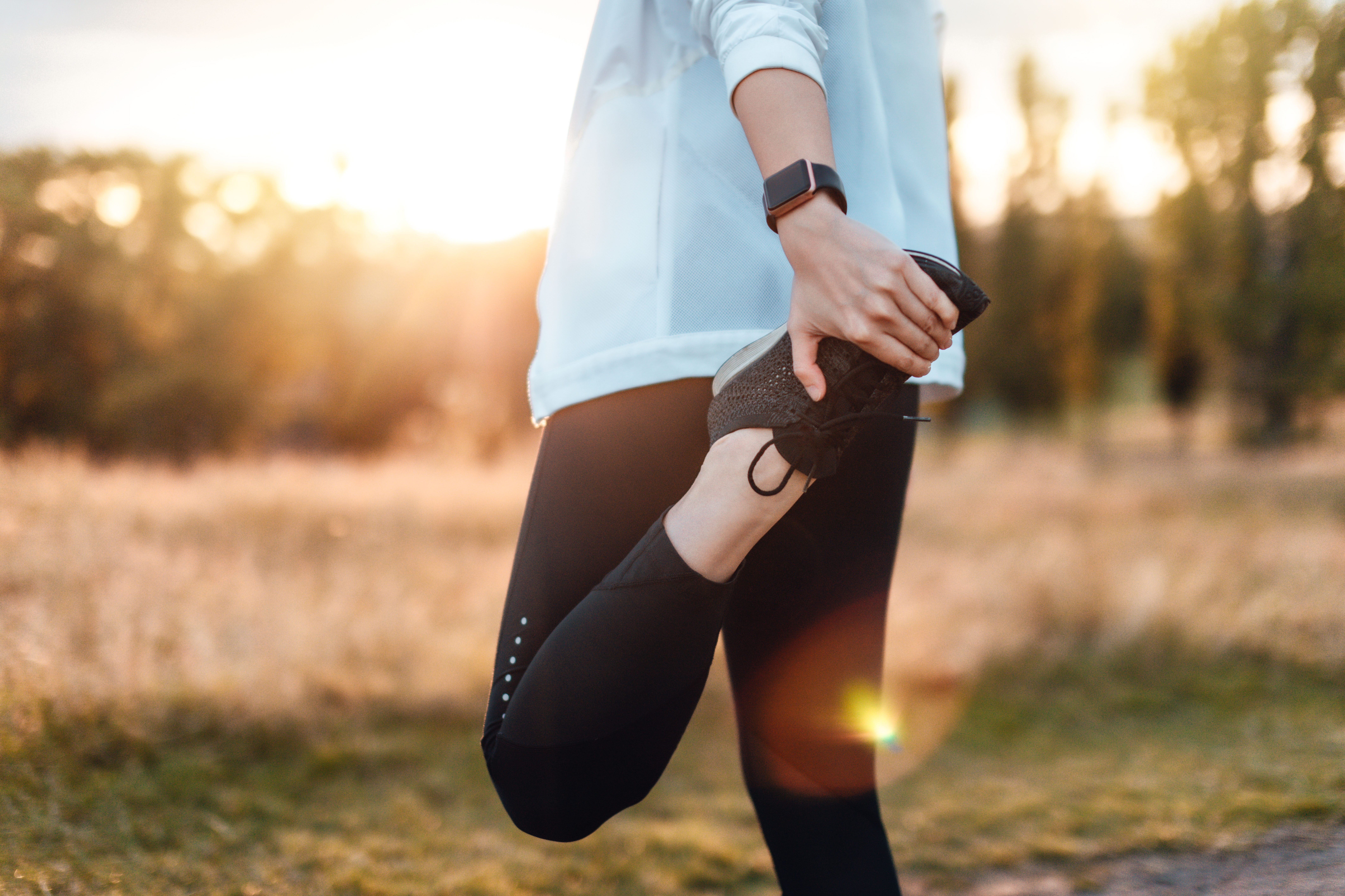 A person wearing athletic clothing stretches one leg by holding their foot behind them outdoors, with sunlight and trees in the background and a fitness watch on their wrist.