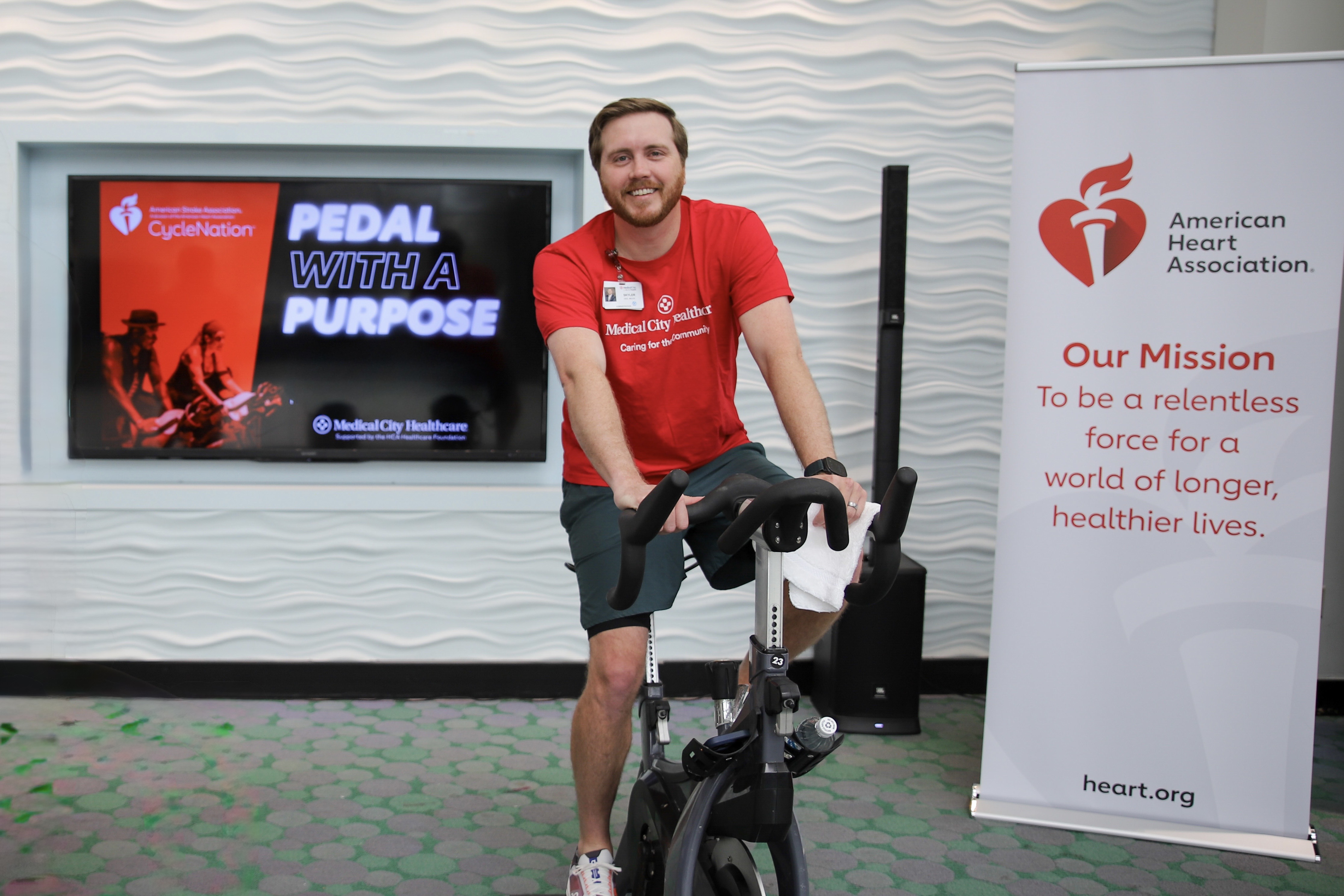 Medical City Children's Hospital CEO Skyler Reed is seated on a stationary bike in front of a screen promoting the CycleNation event