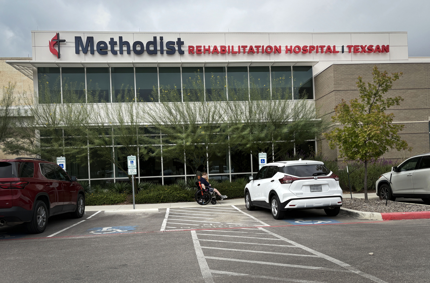 The front entrance of Methodist Rehabilitation Hospital Texsan, showing large glass windows and the facility's name on the building. A person in a wheelchair is visible in the parking lot near accessible parking spaces, with several parked cars in the foreground.