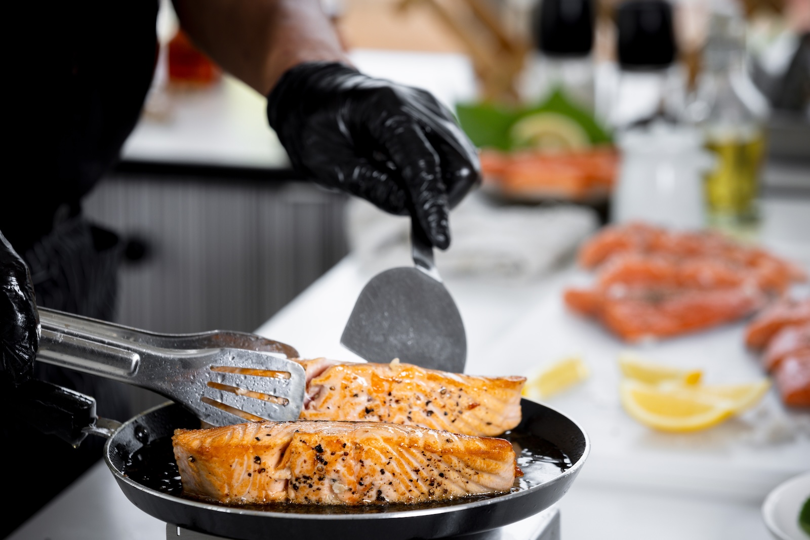 A man cooking two salmon fillets in a frying pan, holding one fillet with tongs and a spatula.