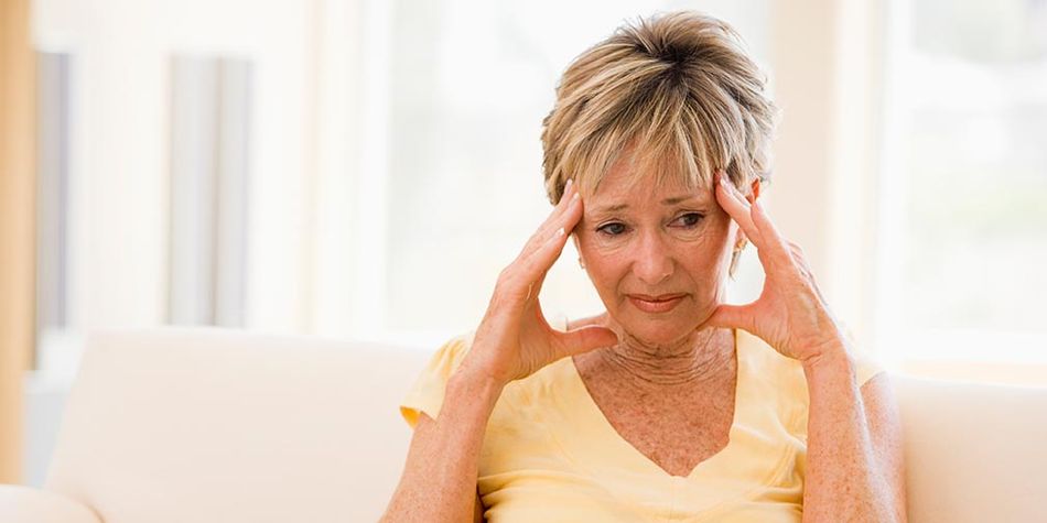 A senior wearing a yellow shirt sits on a white couch, her hands resting on the temples of her head, with a pained expression on her face.