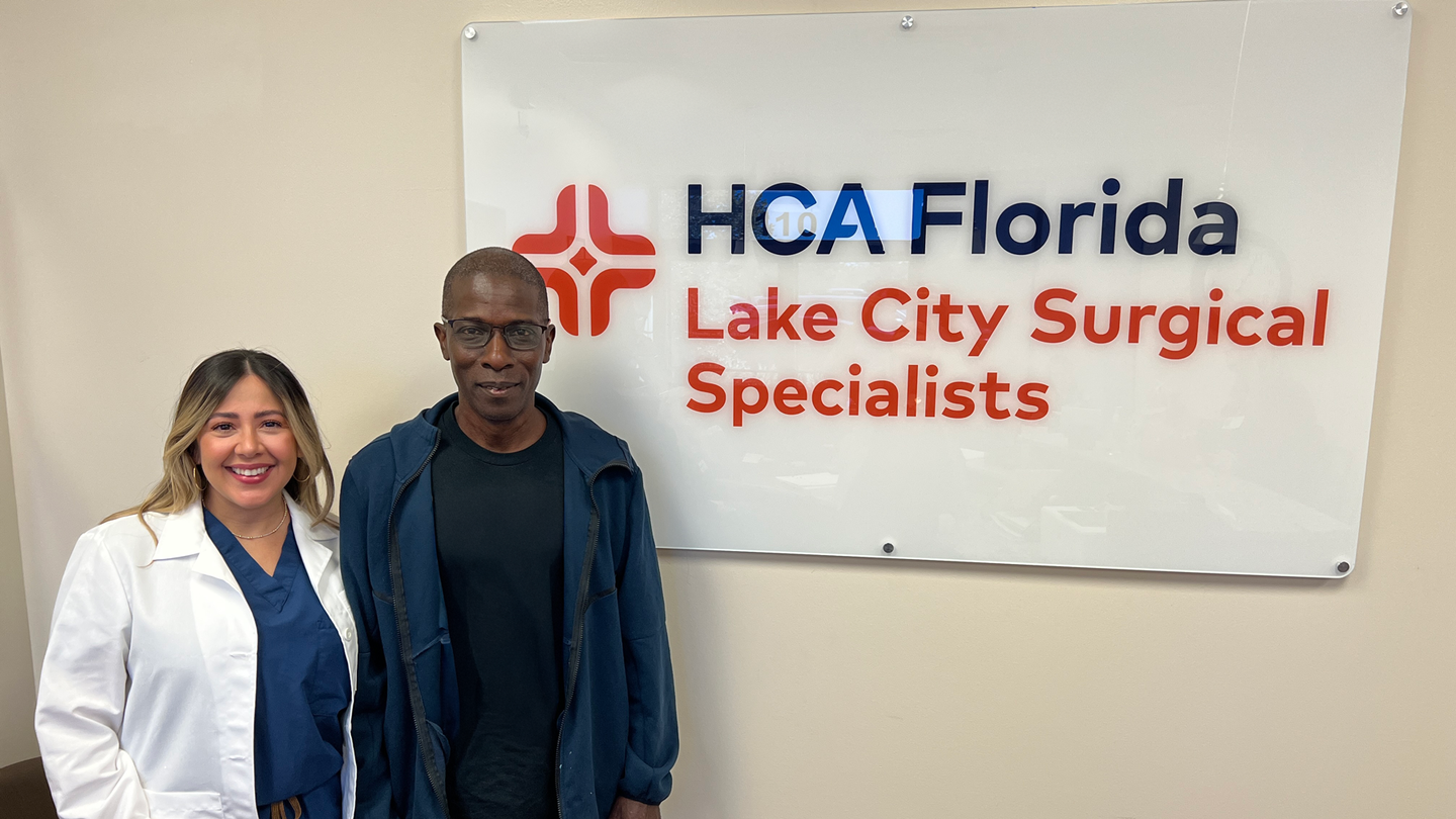 A surgeon with her lab coat on is standing next to a patient in front of the HCA Florida  Lake City Surgical Specialists sign.