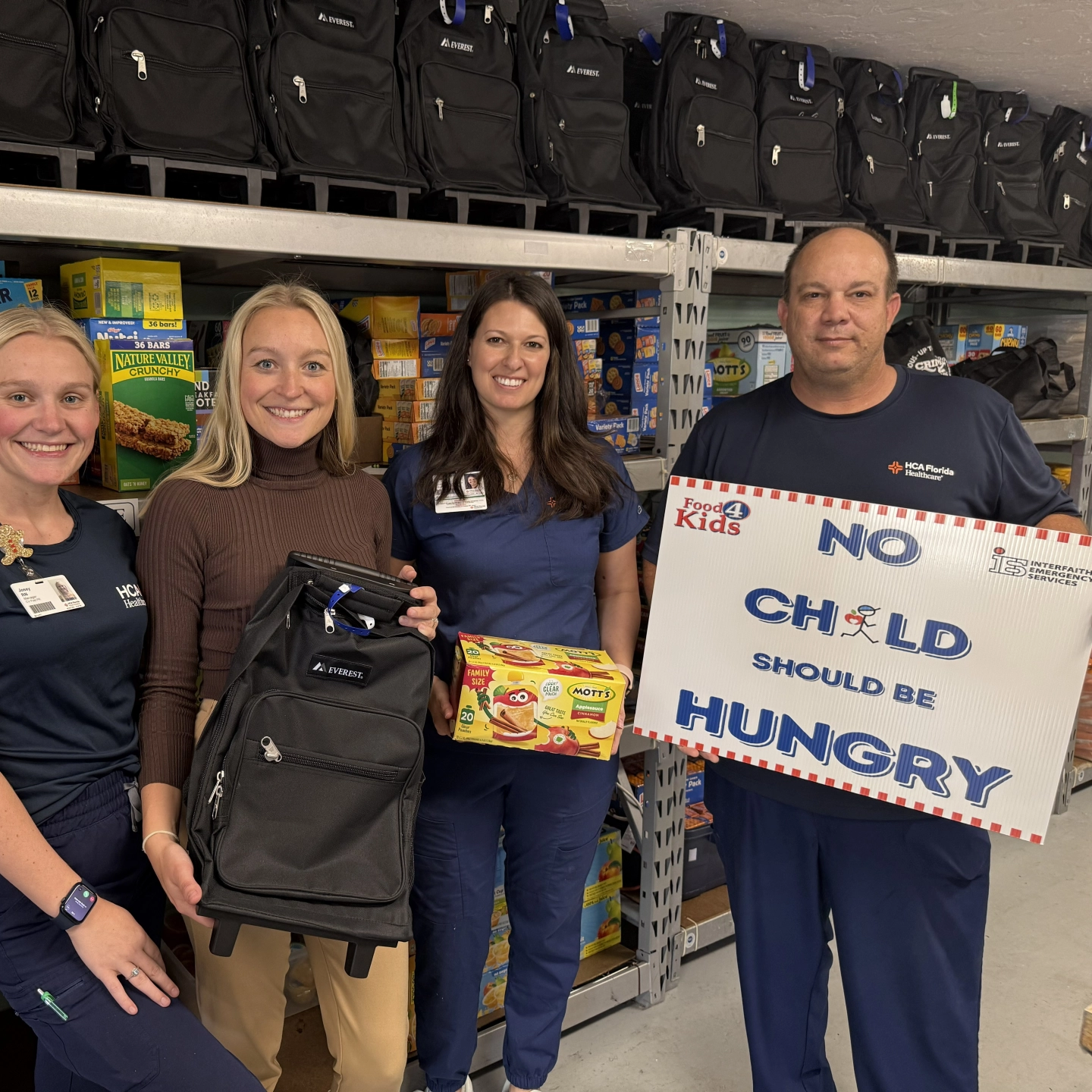 A group of four women and one man most dressed in hospital scrubs, stand in a line smiling and holding a sign that says No Child Should Be Hungry.