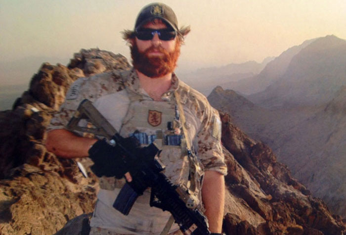 Aaron Kendle, in military gear, standing in mountainous terrain, wearing glasses and holding a rifle.