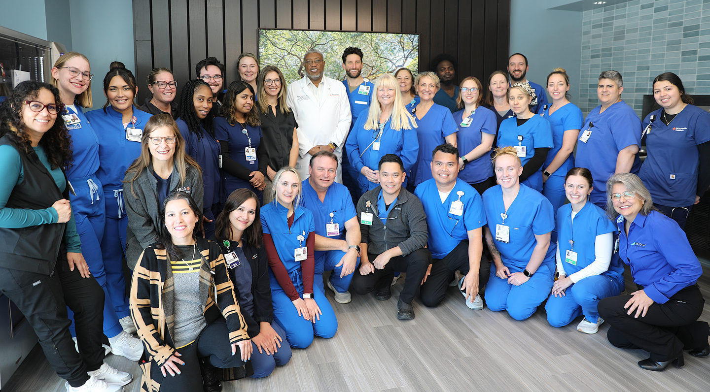 A group of about 30 people, almost all of whom are wearing light blue scrubs, pose in 3 rows in front of a picture of a large tree.