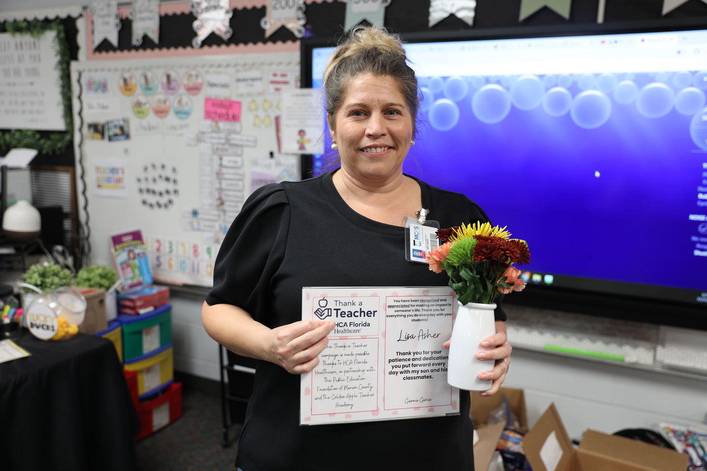 Shows a woman in a black t-shirt smiling as she holds a certificate and a small bouquet of flowers.