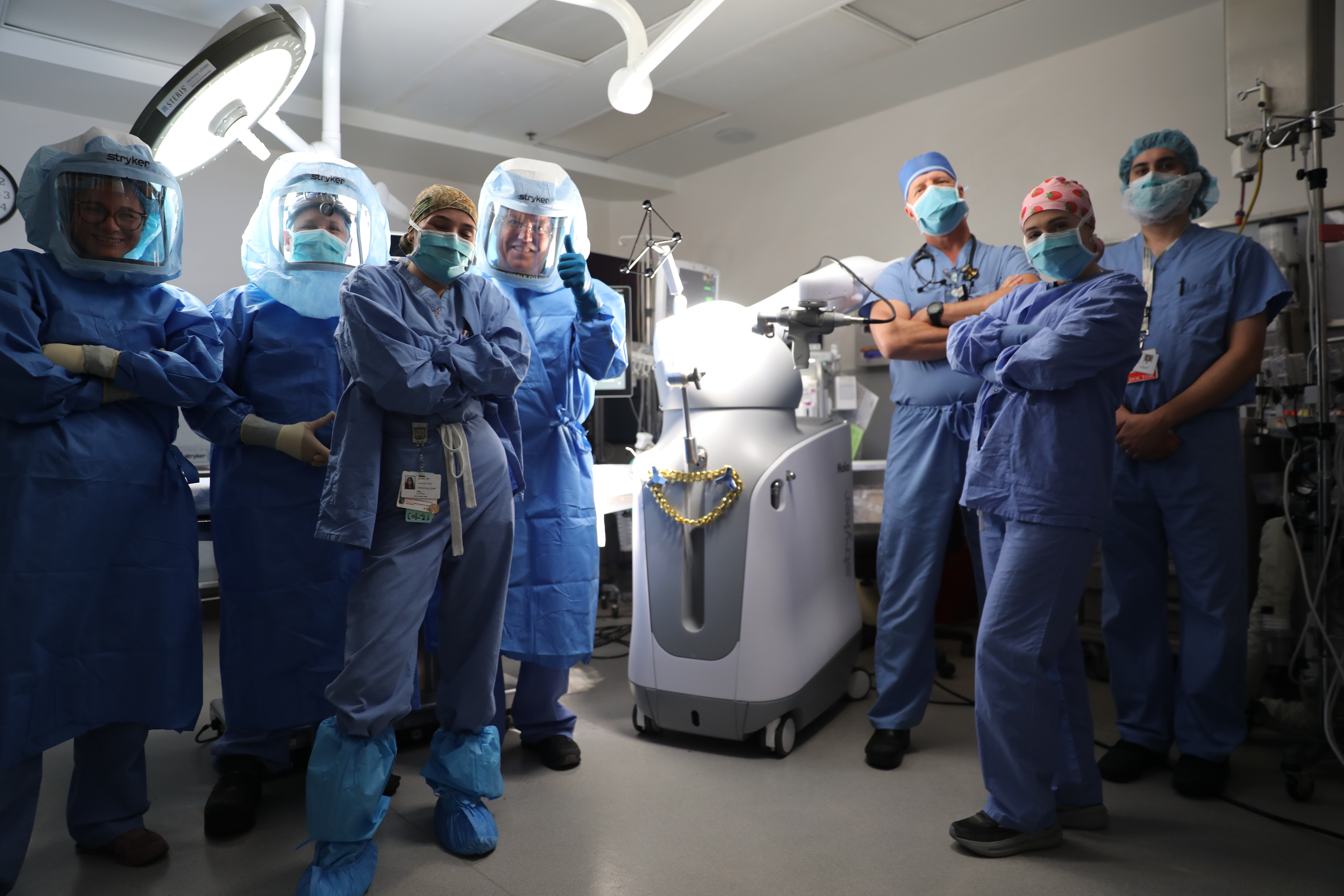 A group of doctors and nurses in hospital scrubs and masks stand in an operating room around a surgery robot