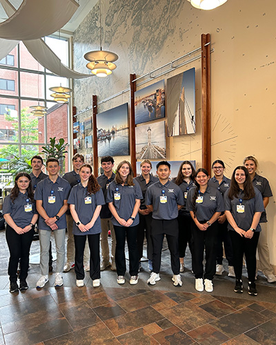 Portsmouth Regional Hospital Summer Healthcare Exploration Program volunteers pose in two lines of seven people wearing their grey program t-shirts and badges inside the hospital.