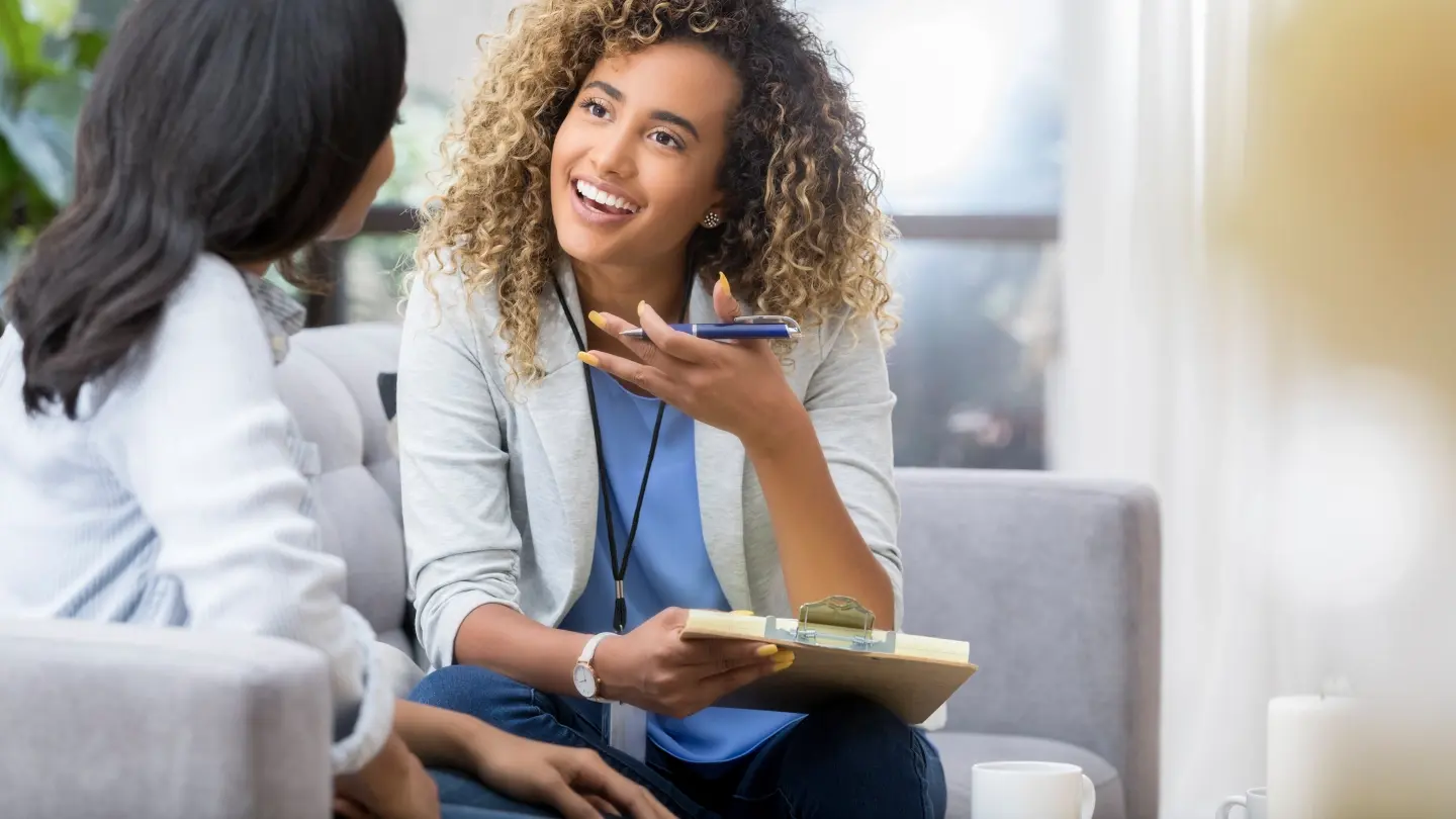 A female therapist gestures as she talks with a female client while sitting on a couch.