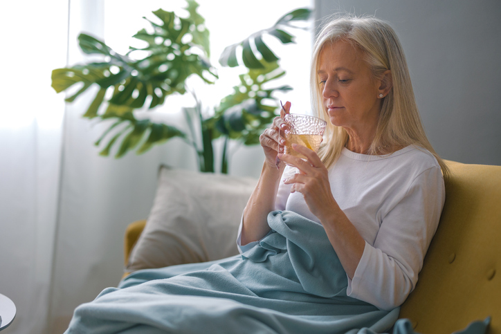 A mature woman sitting on a couch at home under a blanket, drinking tea.