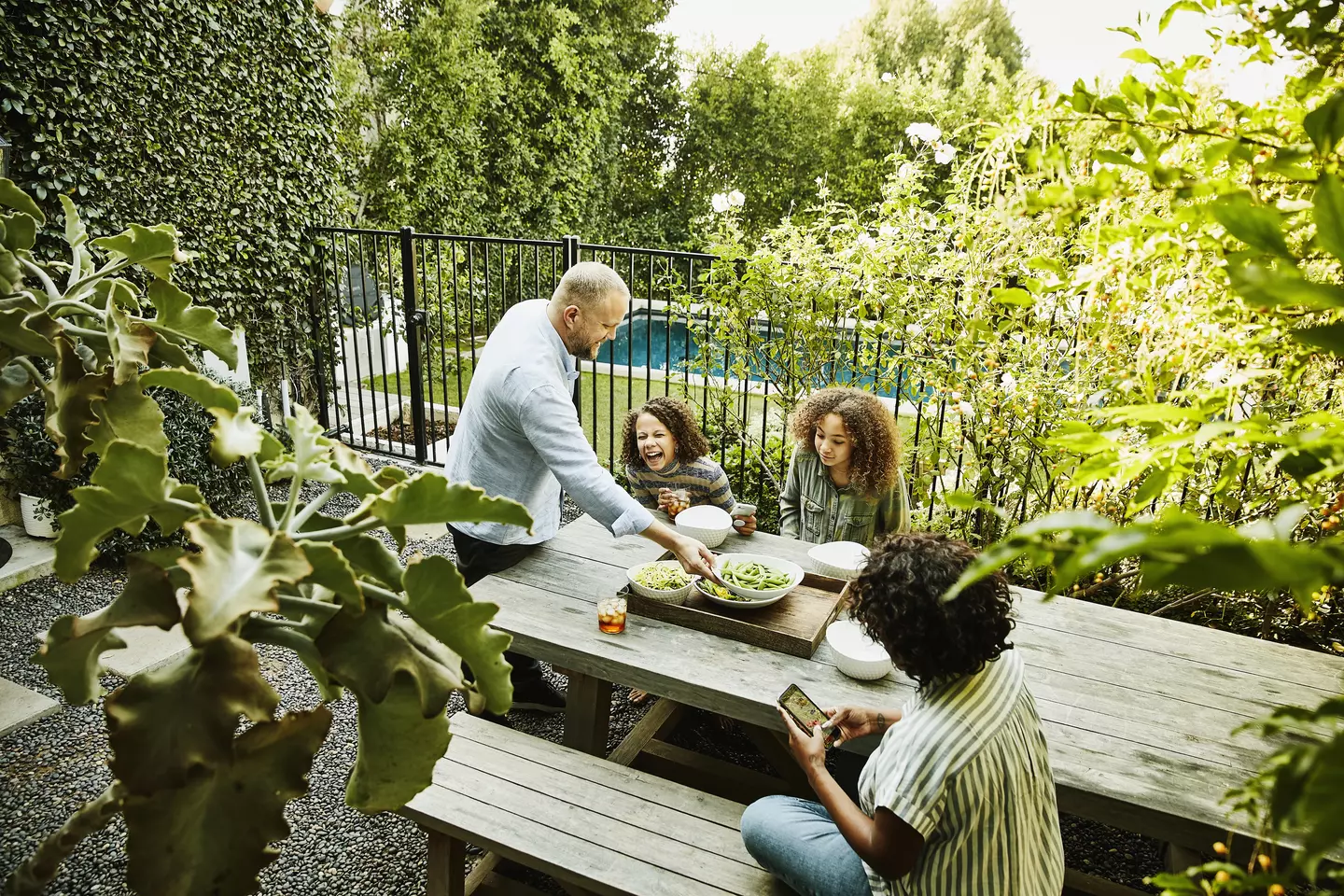 A smiling and laughing family of four shares a meal at a picnic table in a courtyard outside, with a swimming pool in the background.