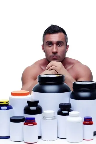 Shirtless man sitting behind protein and supplement bottles.