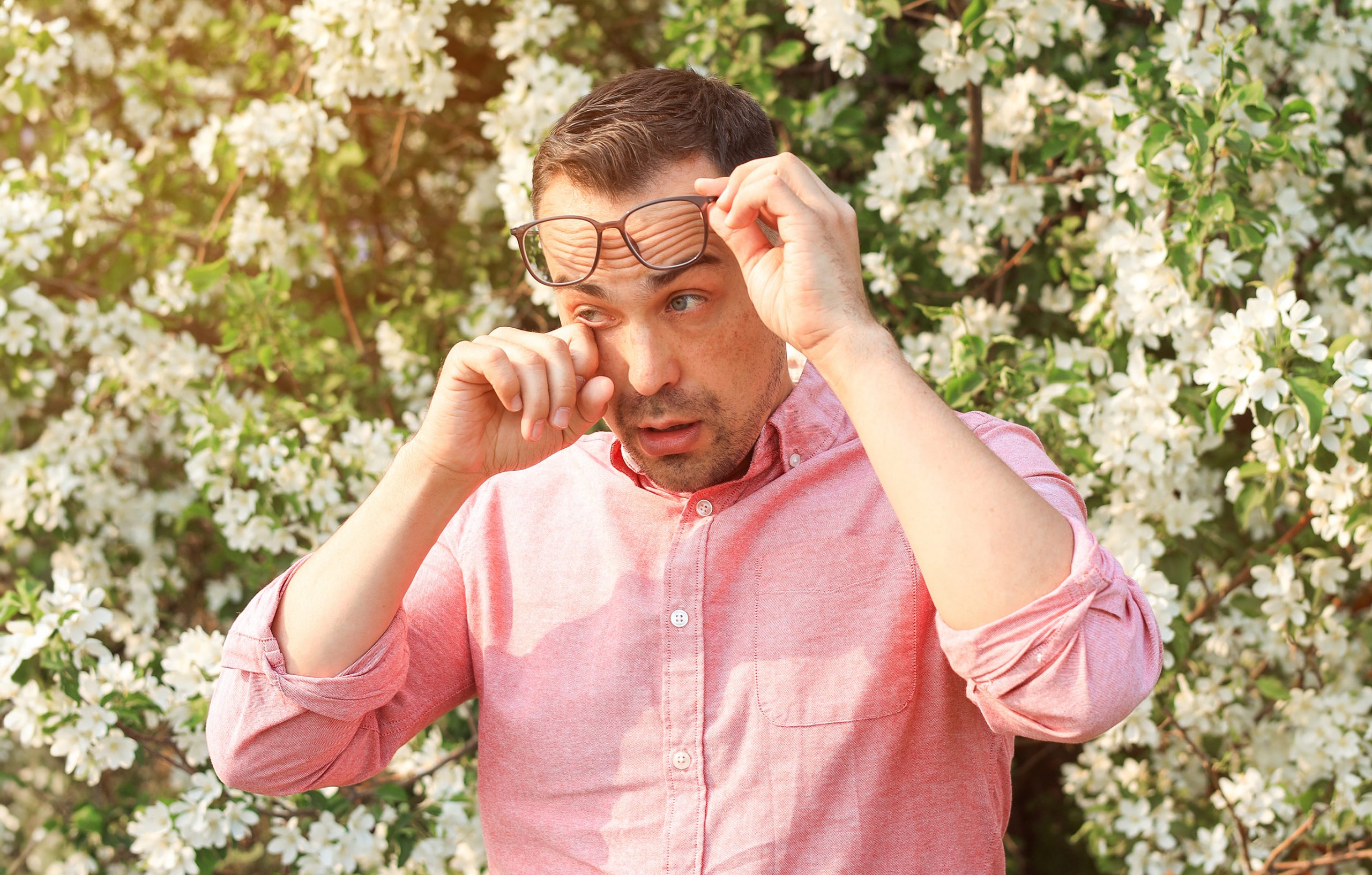 A man in a pink button-down shirt raises his eyeglasses to wipe his eyes, while standing in front of flowers.