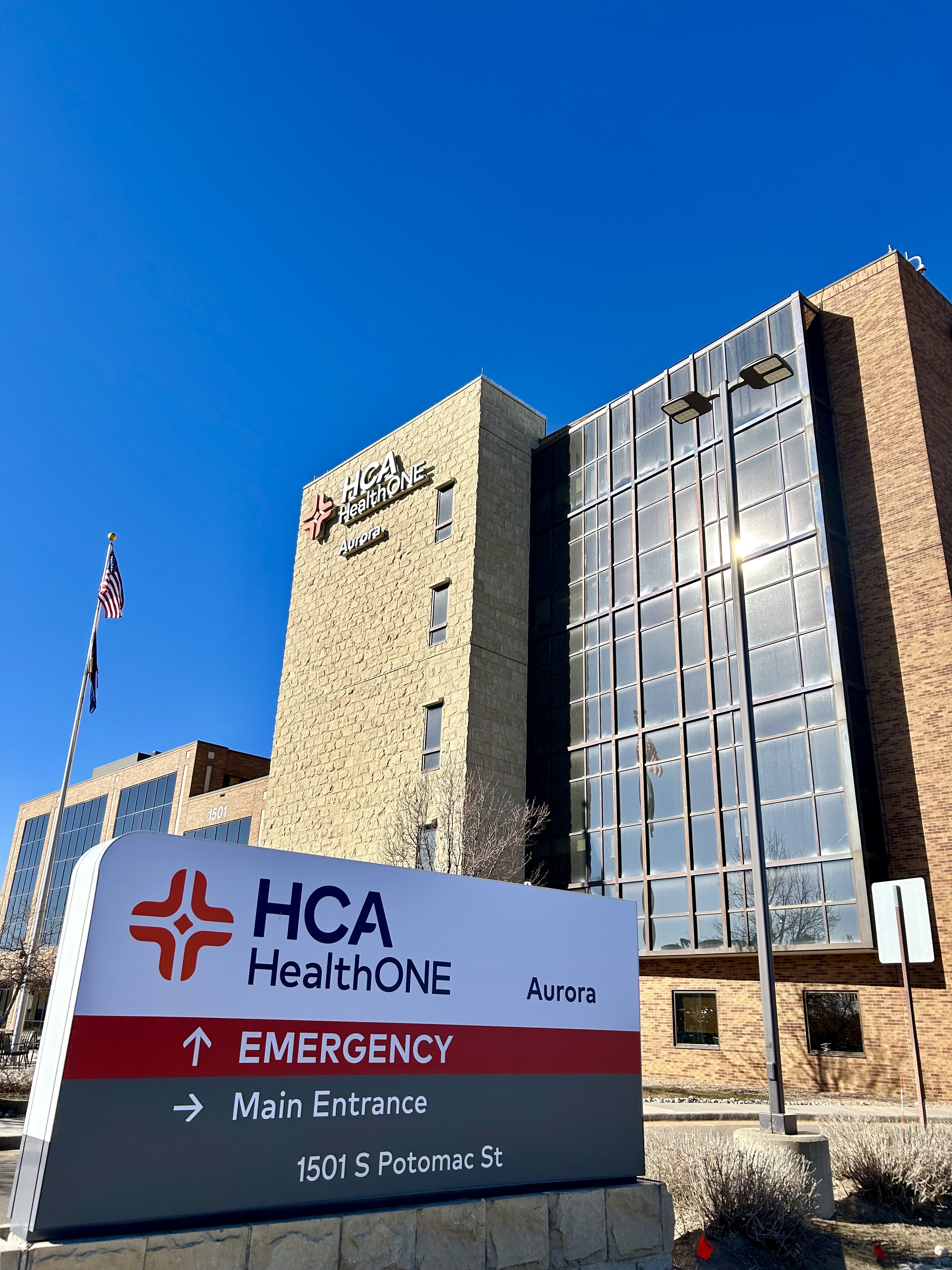 Entrance sign in front of HCA HelathONE Aurora. White, red, and gray sign directing towards Emergency and Main Entrances. A tall multi-story brick building with black glass panneled windows.