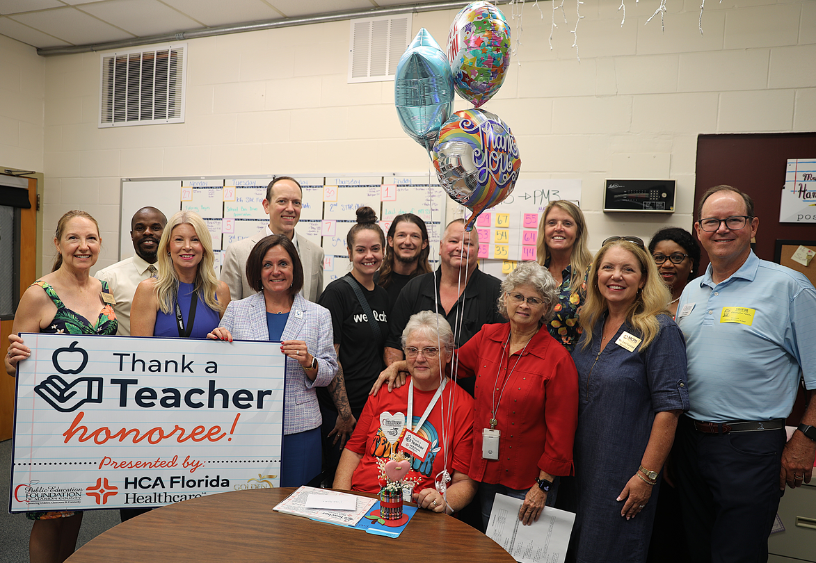 A group of educators, community leaders, and healthcare representatives gather in a classroom to celebrate a teacher recognition award. Several people stand behind an honored teacher seated at a table, while others hold a large "Thank a Teacher Honoree!" sign presented by HCA Florida Healthcare and local education partners. Colorful balloons and classroom schedules decorate the room.