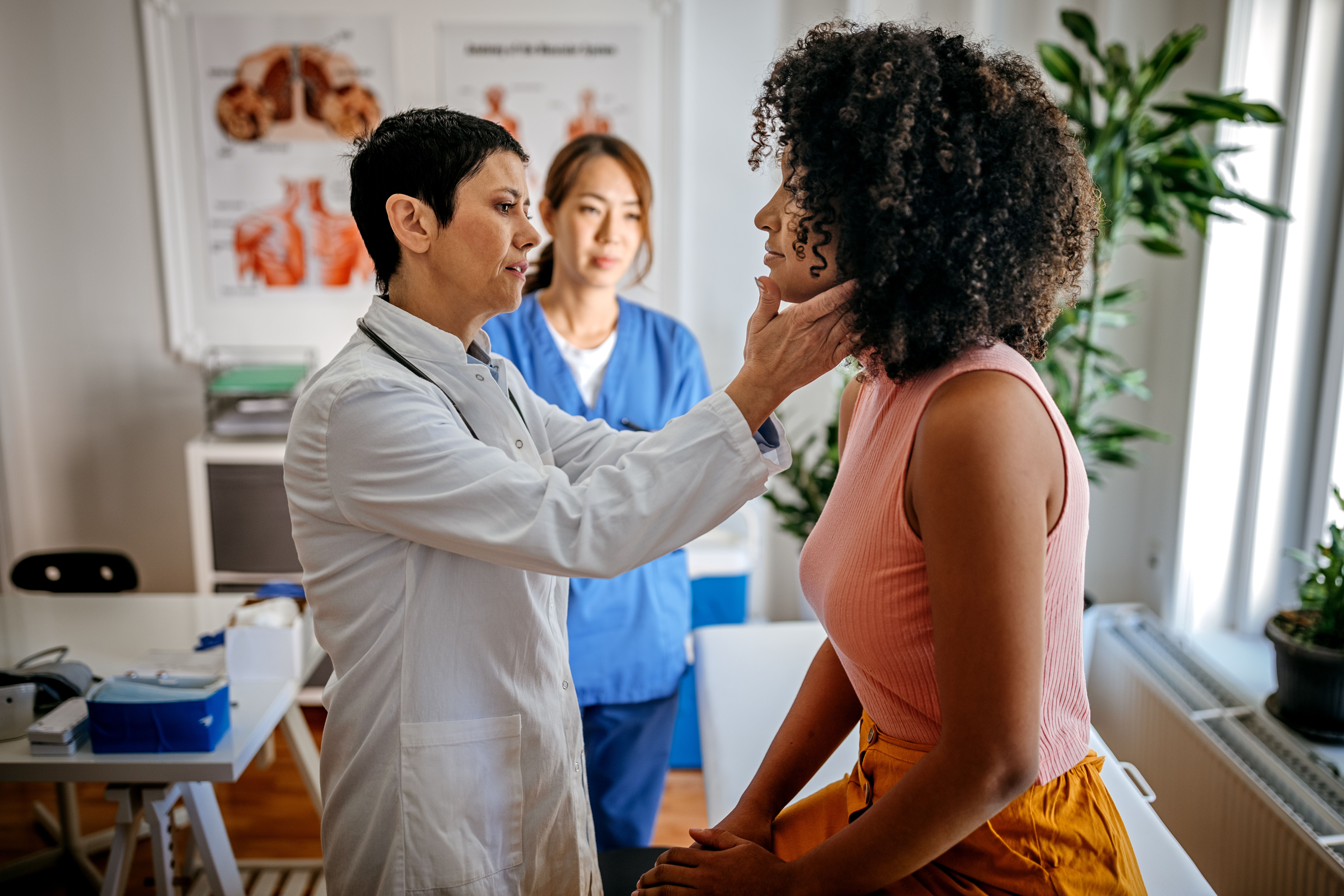 A doctors checks a woman's thyroid in a patient exam room with an attendant behind them.