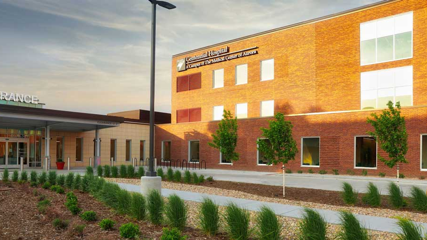 The exterior view of Centennial Hospital displays the brick building partially sunlit, with a shaded tree and plant lined landscaped walkway leading to the entrance
