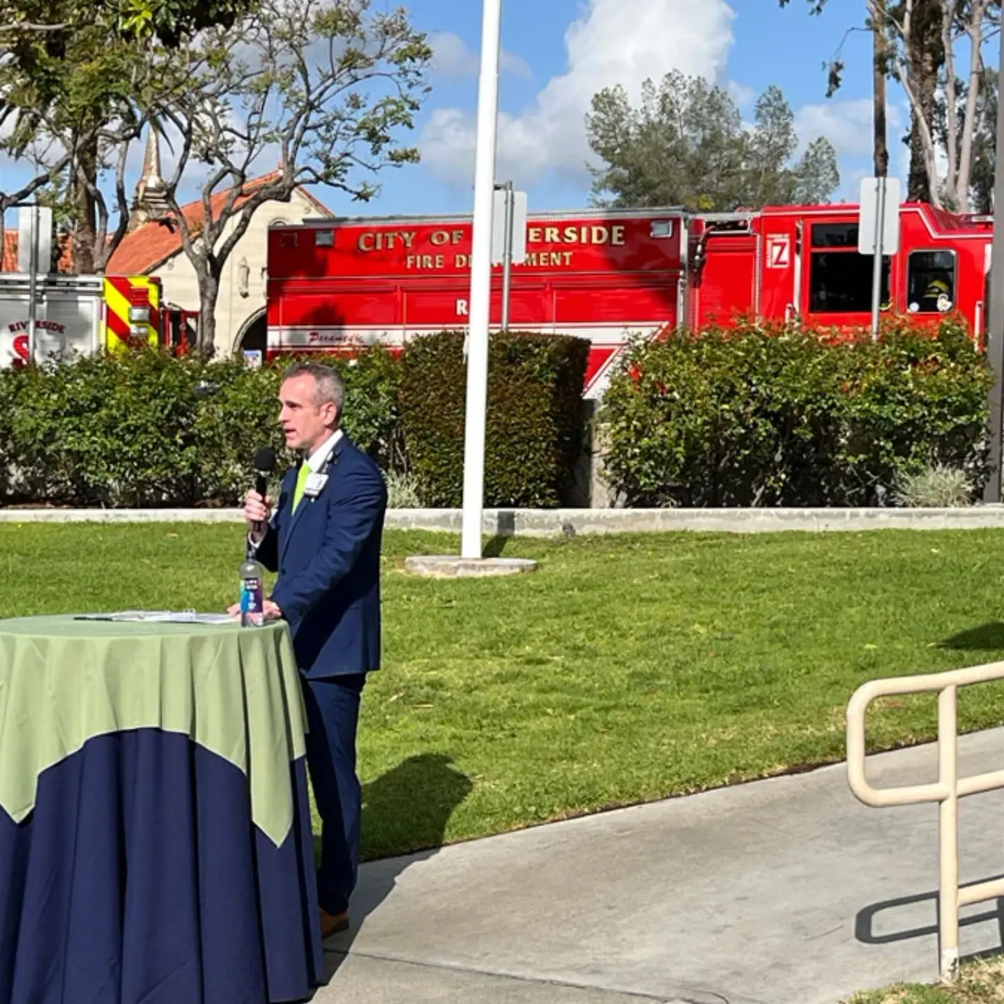 Peter Hemstead, CEO of Riverside Community Hospital, talks at a standing table with emergency vehicles in the background.