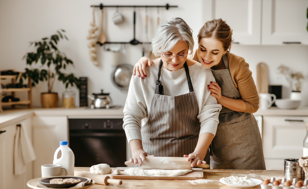 Mother and adult daughter rolling out dough with rolling pin on wooden table.