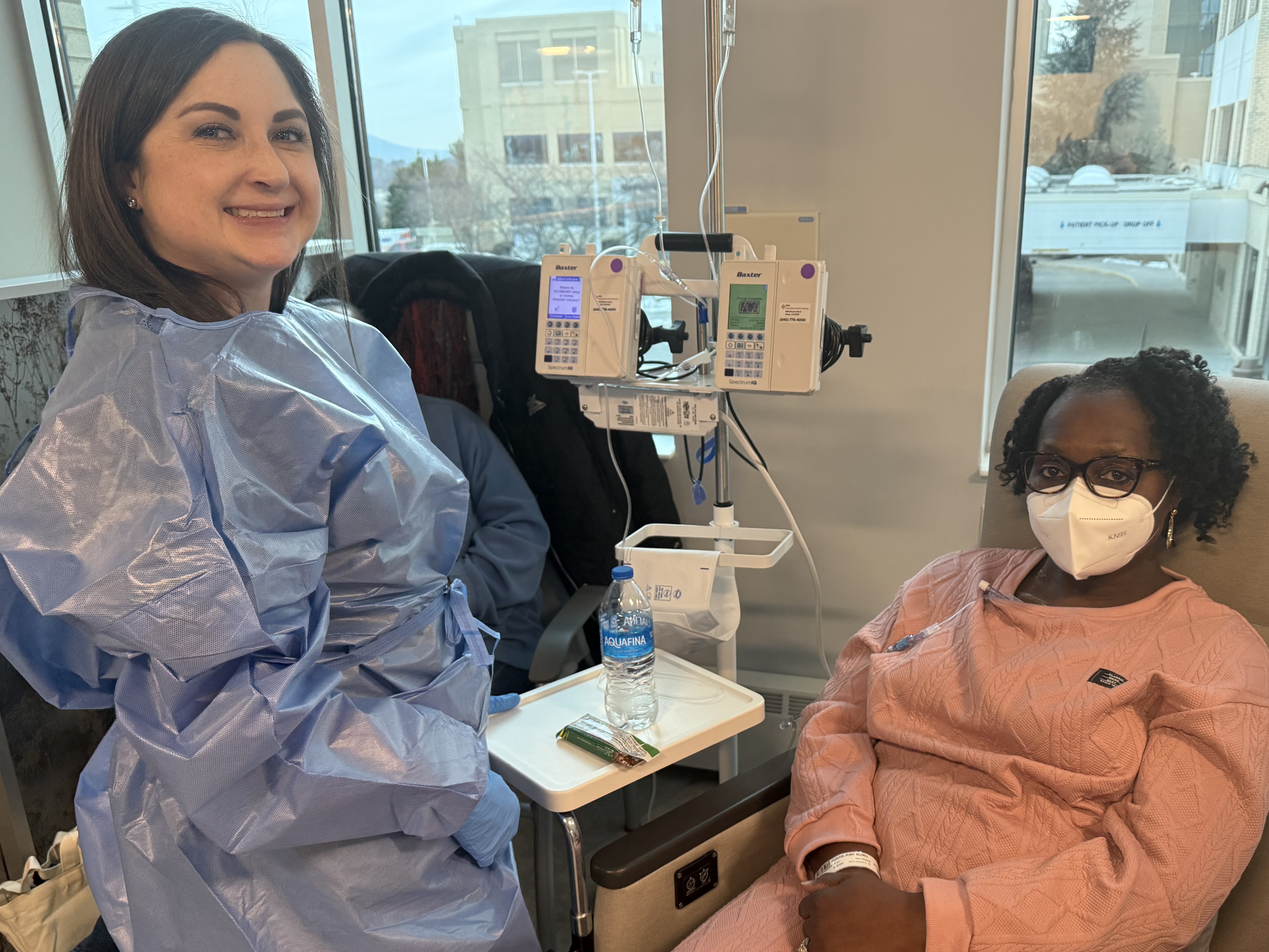 Patient Penni Hairston is seated in a chair receiving an IV cancer treatment. Her nurse, Kayla, is standing next to her wearing medical scrubs.