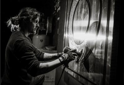A black and white image of Eric Breish, wearing his hair in a bun, sanding one of his commissioned pieces.