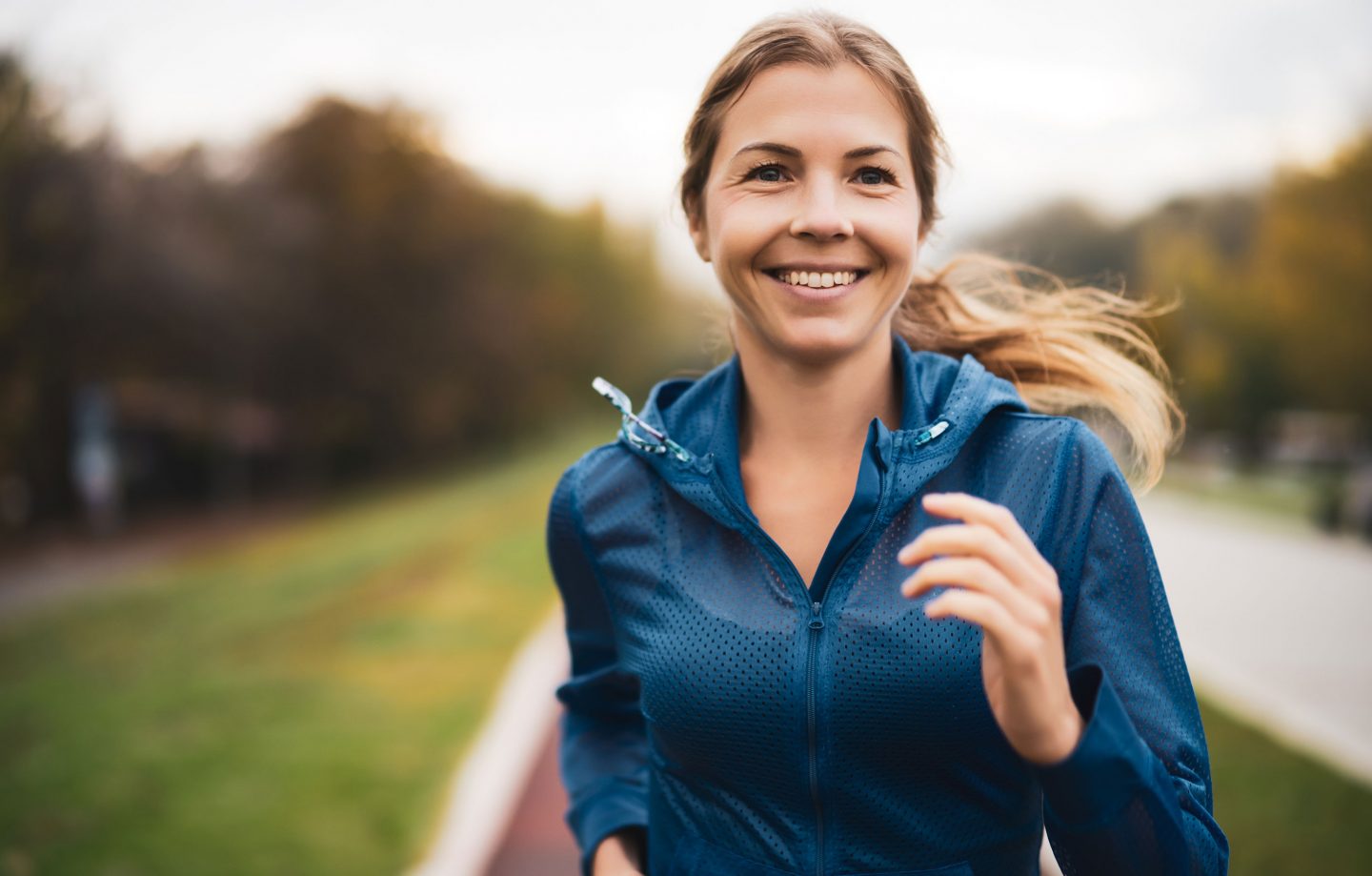 A woman smiles while jogging, wearing a light windbreaker jacket.