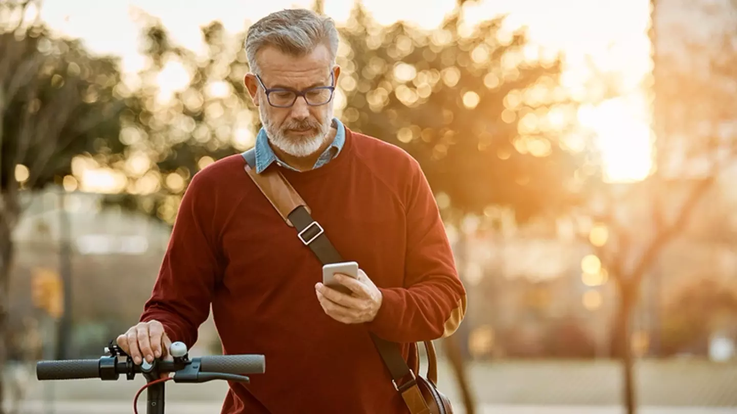 An older man looking at his smartphone while walking with a scooter bike in the city, as the sun sets behind him.