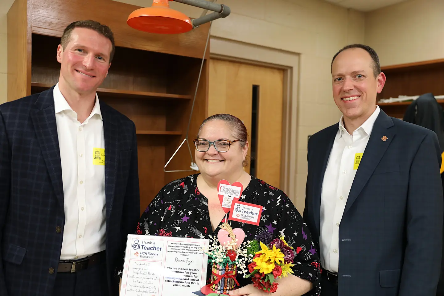 A woman holding a bouquet of flowers and certificate stands between two men and all of them are smiling.