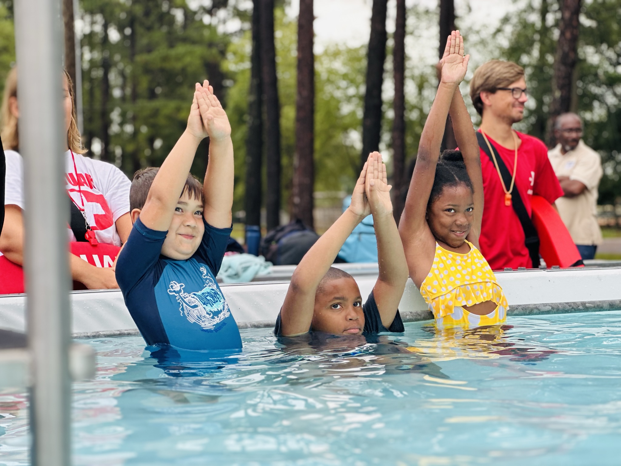 Three children in a swimming pool, standing with their arms raised in a diving position, learning how to swim.