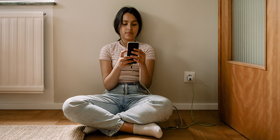 A girl using smart phone while sitting cross-legged on floor against wall at ho.me