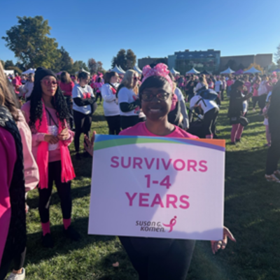 Woman in a pink shirt and headband smiles while holding a large sign that reads "SURVIVORS 1–4 YEARS – Susan G. Komen" at an outdoor breast cancer walk, surrounded by a crowd of participants wearing pink.
