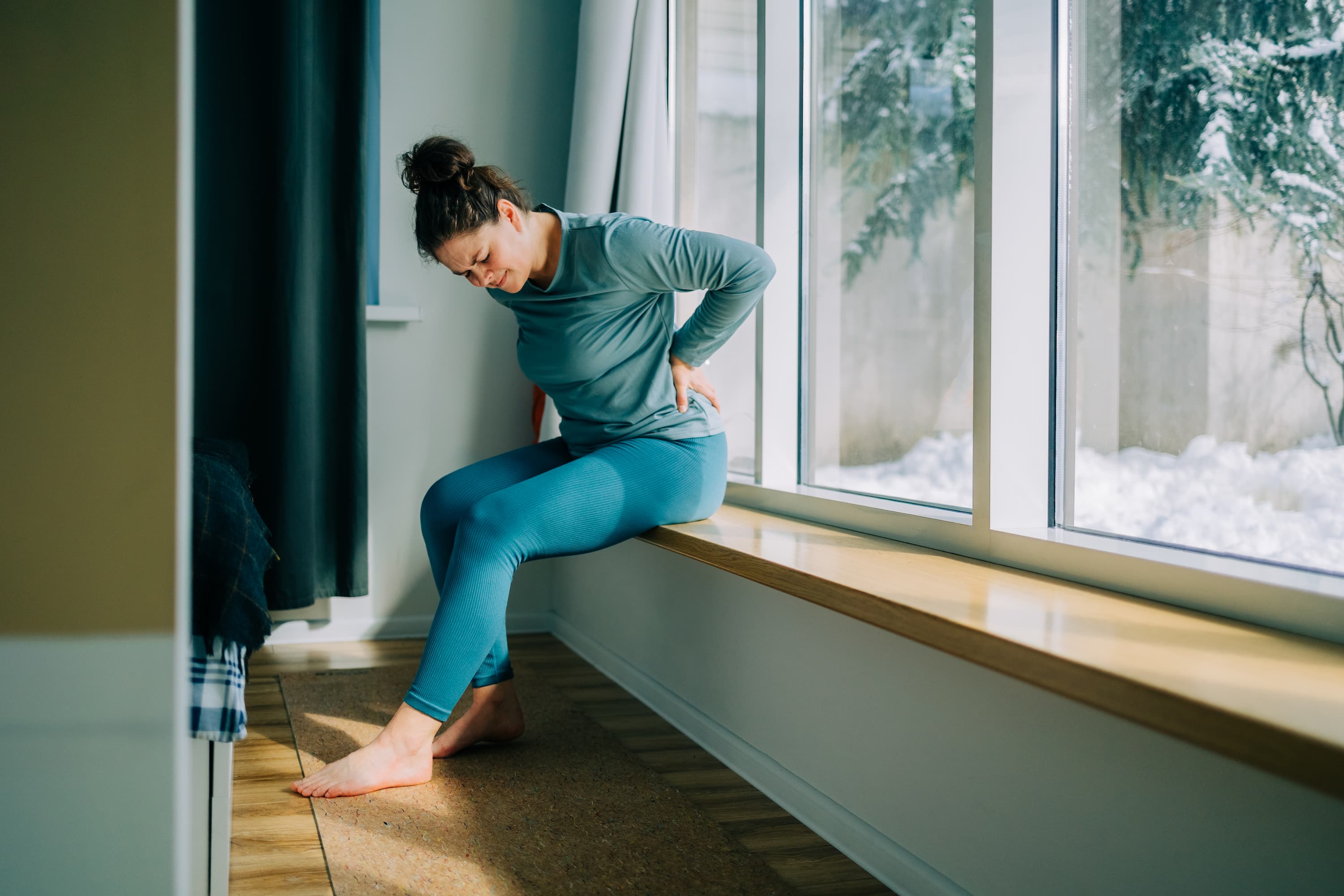 Woman sitting on a window ledge at home, holding her lower back in pain.