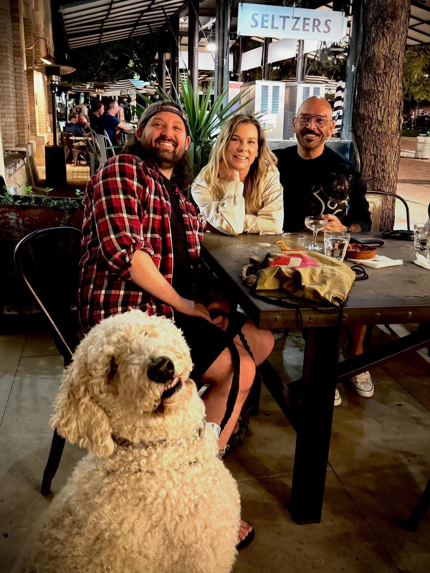 Rick Gipprich and two friends sit together at an outdoor restaurant table in the evening, surrounded by warm string lights and a casual patio atmosphere.