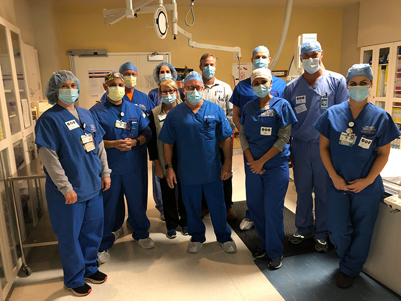 Dr. Charles Machell and his cardiovascular team, wearing masks and scrubs, gather together in a surgical room for a photo.