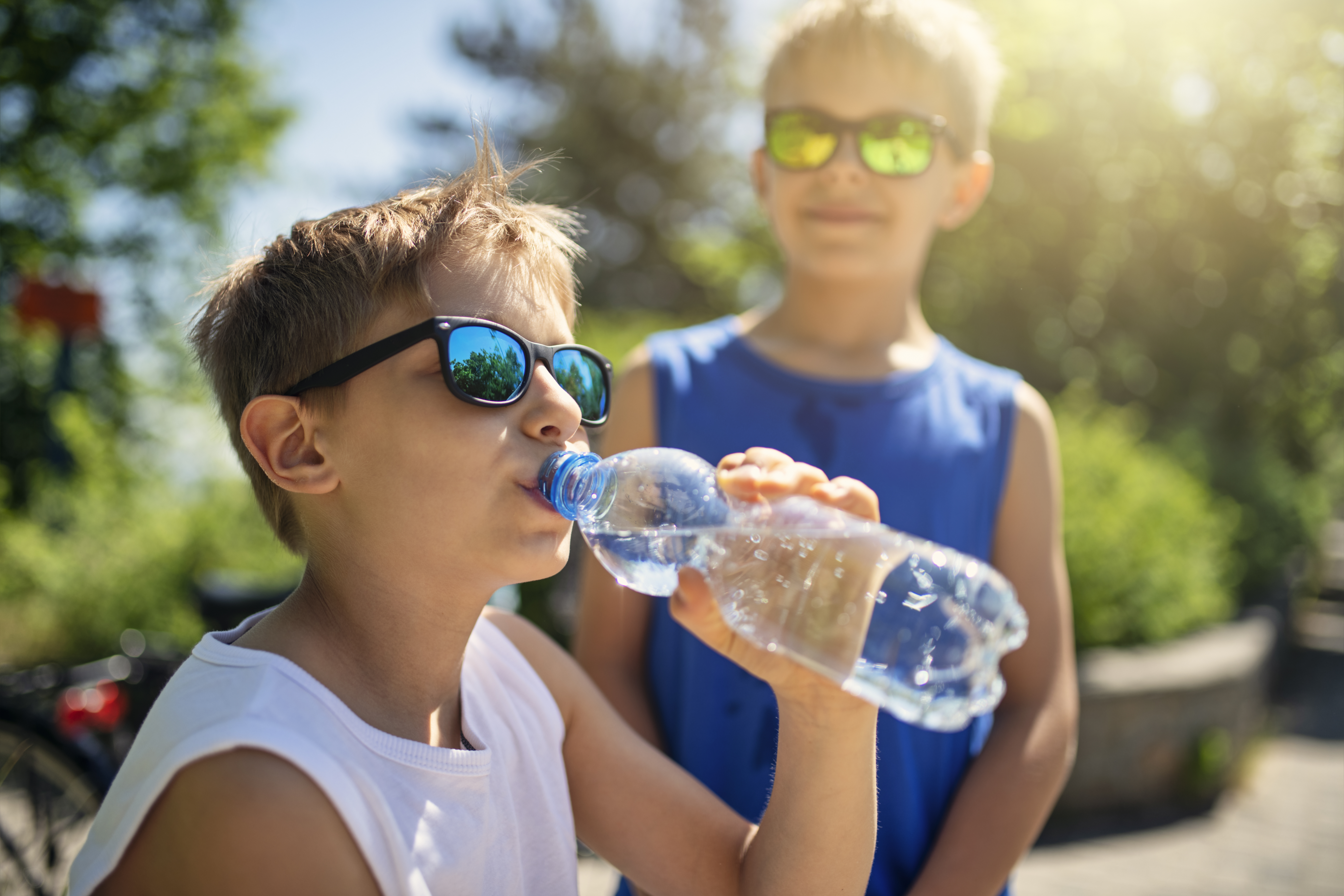 A young boy wearing sunglasses drinks from a plastic water bottle, with another boy in sunglasses standing behind him.