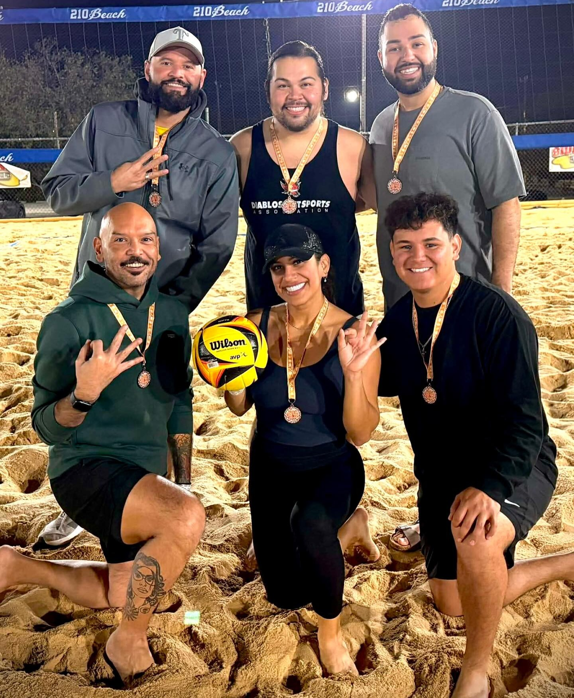 Rick&nbsp;Gipprich&nbsp;and friends pose together on a sand volleyball court at night, wearing matching medals and holding a yellow Wilson volleyball. They are kneeling and standing in a celebratory pose, with the court’s net and lights visible in the background