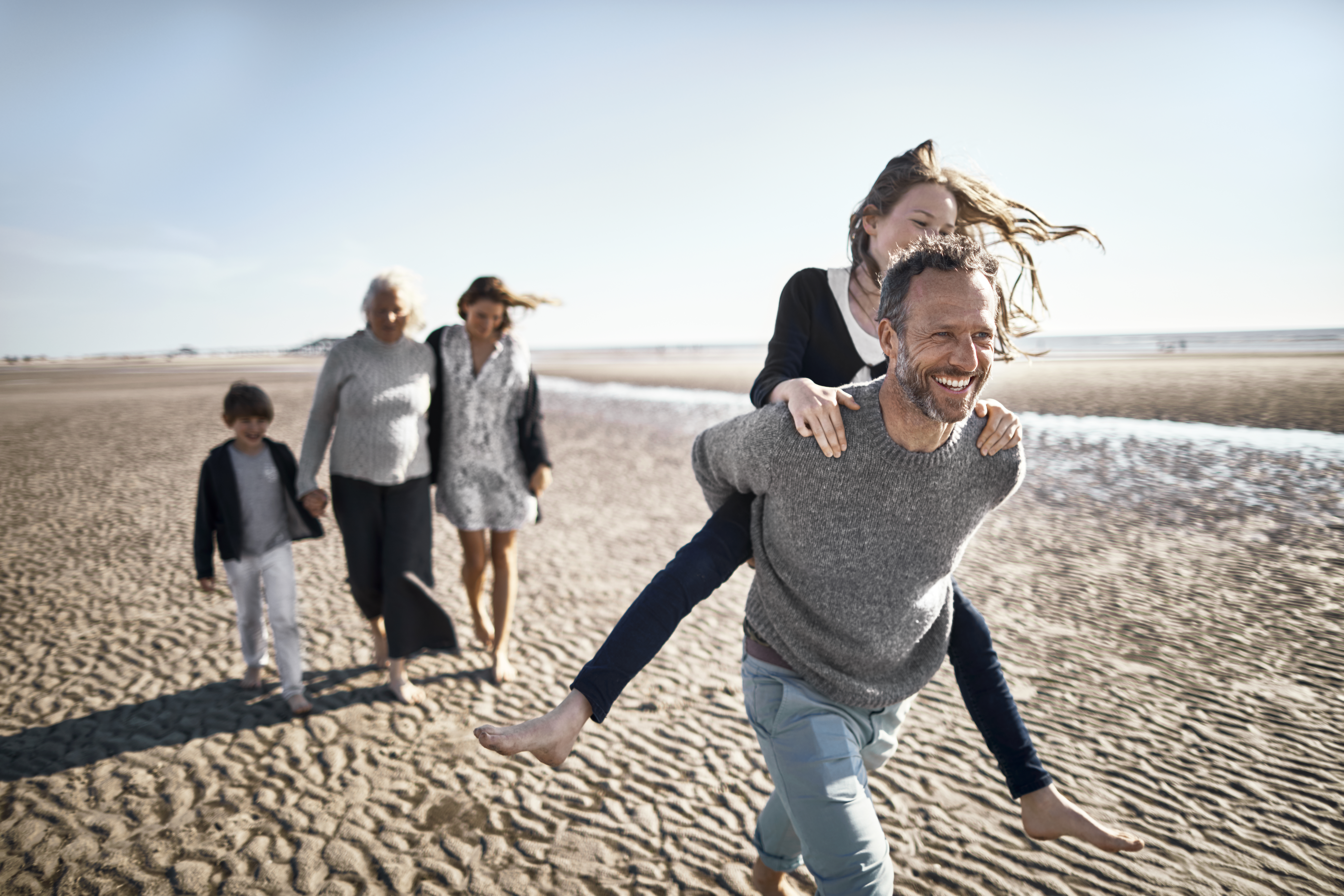 A smiling father carries his daughter on his back across a beach, while family members follow behind them.