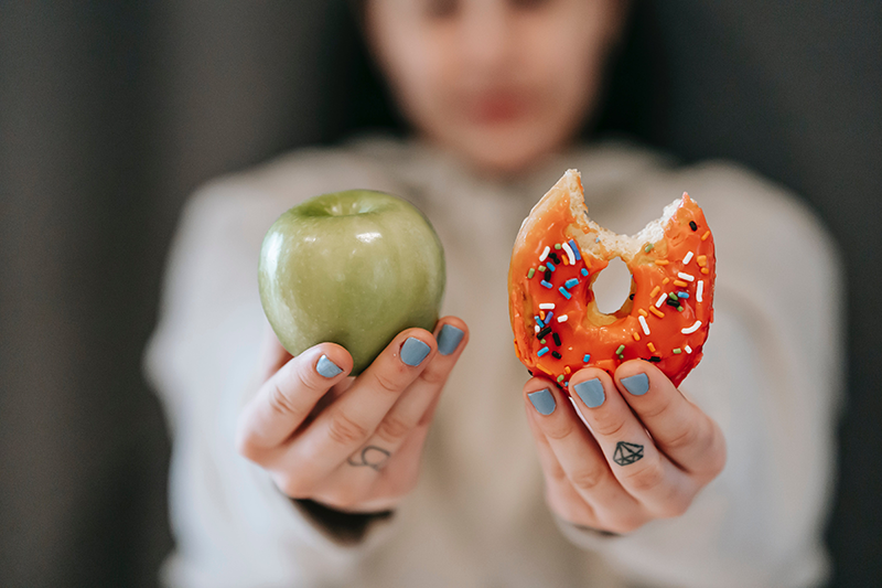 A woman with her arms outstretched, holding a green apple in one hand and a donut with sprinkles in the other.