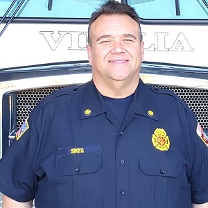 Brian Sikes standing in front of a fire truck, wearing his fireman's uniform.