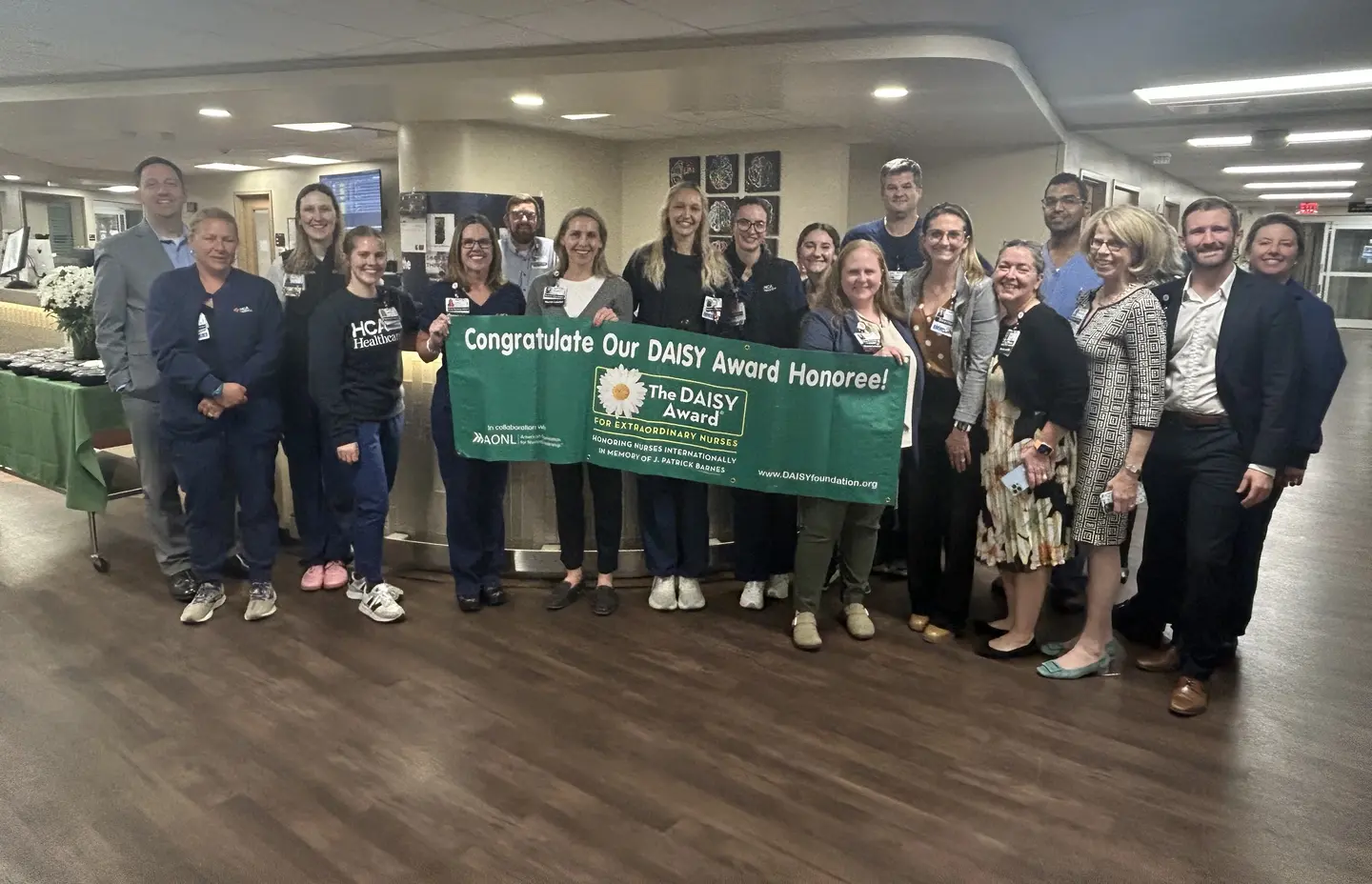 Bailey DiNucci and a dozen of her team posing with DAISY award banner in hospital lobby.