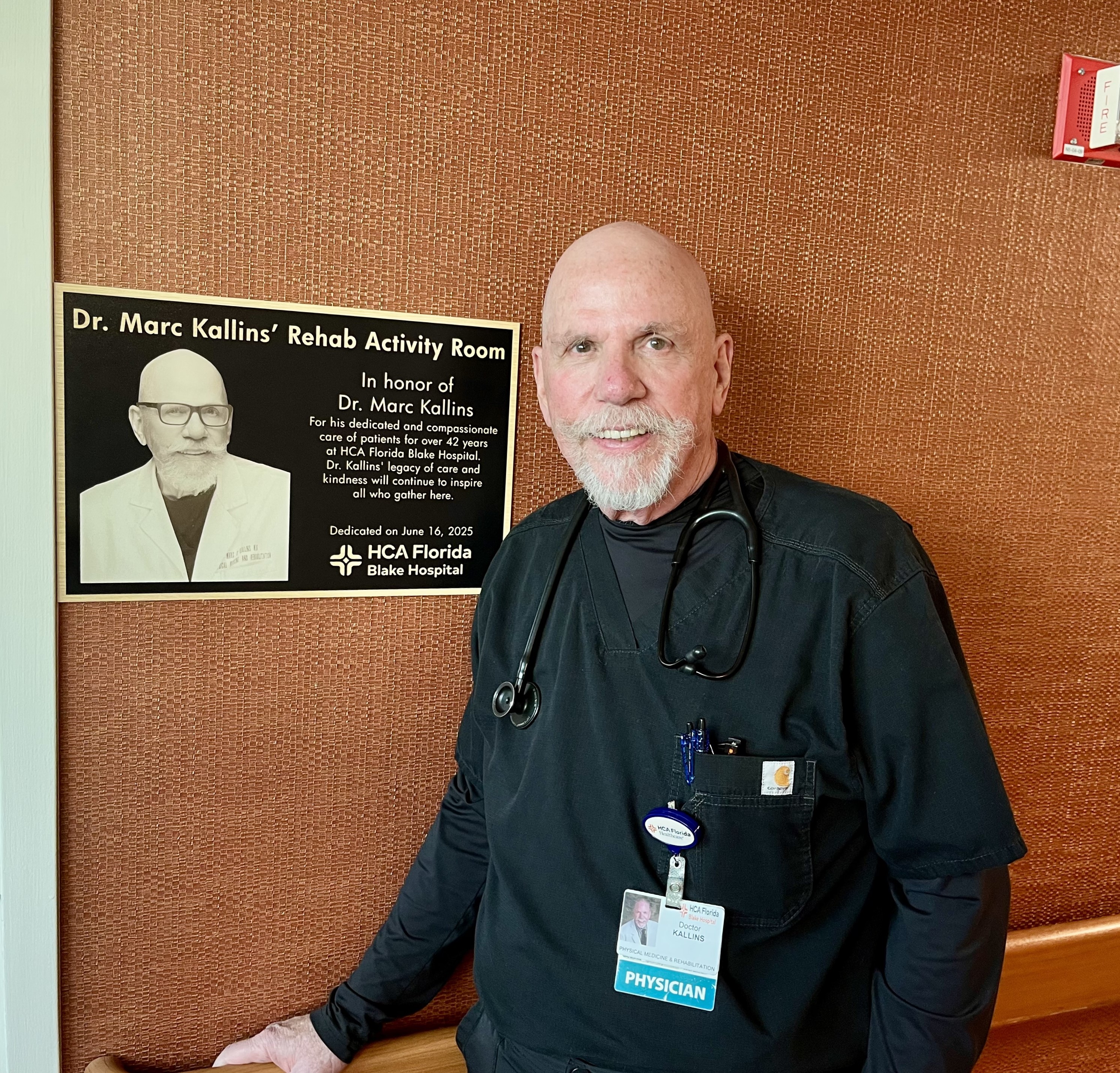 Dr. Marc Kallins stands beside a commemorative plaque that officially renamed the Coquina Room to the Dr. Marc Kallins Rehab Activity Room.