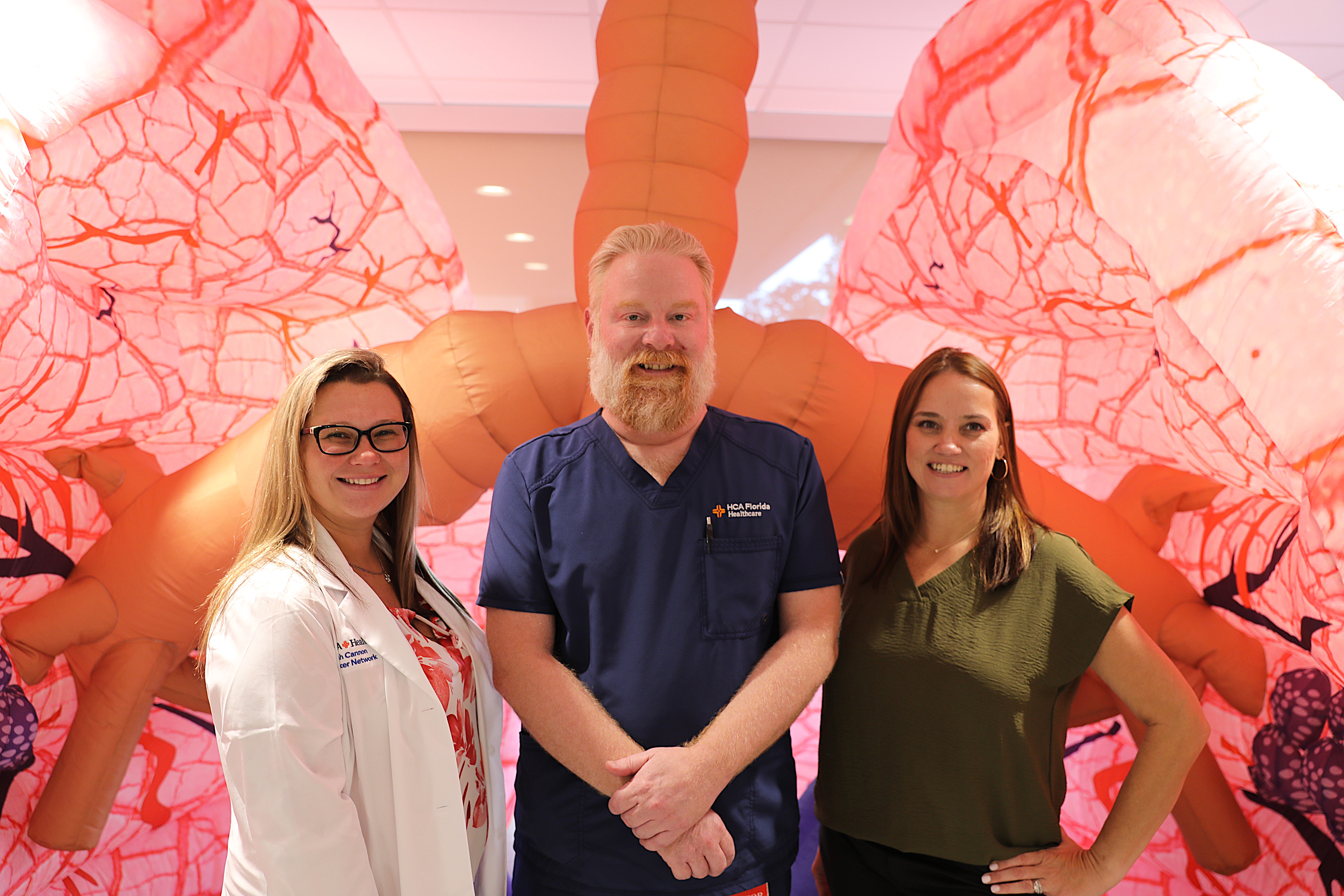 The picture shows a man in blue scrubs and two women standing in front of a pink-and-red set of inflatable lungs.
