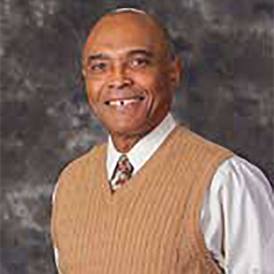 Louis Sylvester smiles while wearing a white button-up shirt and brown sweater vest.