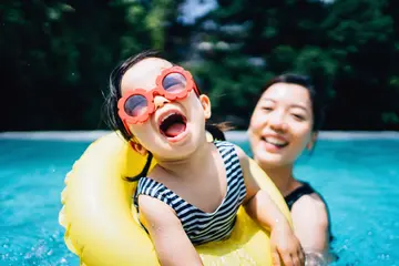 A toddler wearing flower shaped sunglasses plays in an outdoor swimming pool with her mother.