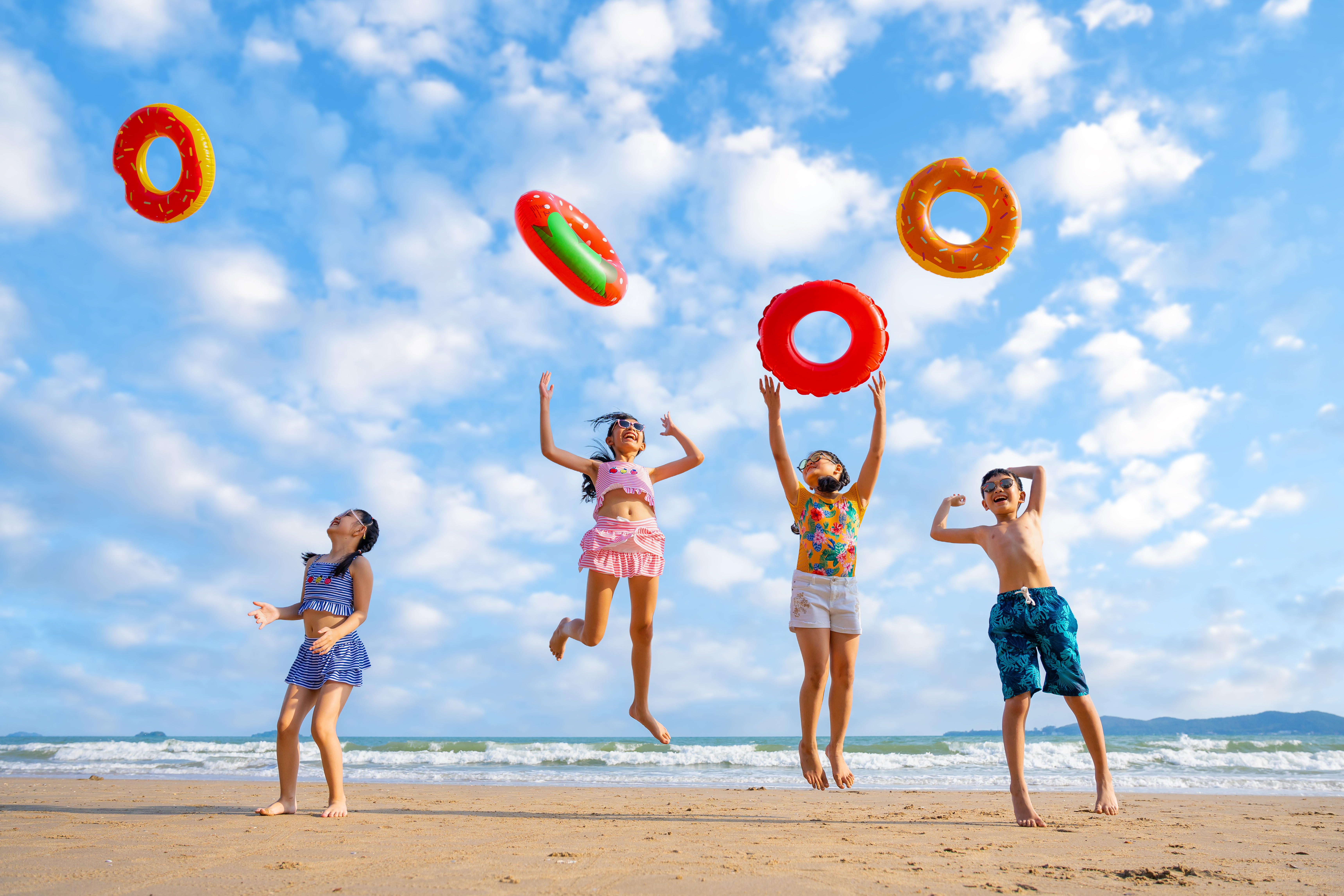 Children play on a sunny beach, jumping and tossing inflatable swim rings near the ocean.