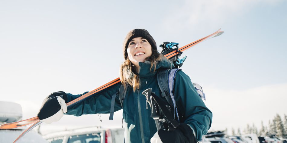A smiling woman carrying skis during winter, with cars and trees in the background.