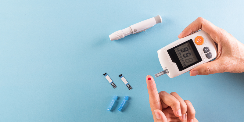 Against a blue backdrop, a hand holds a blood glucose meter next to a drop of blood on a finger, alongside a lancing device, lancets, and test strips.