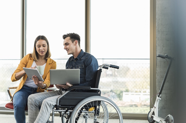 Young man with laptop in wheelchair and woman with tablet in office.