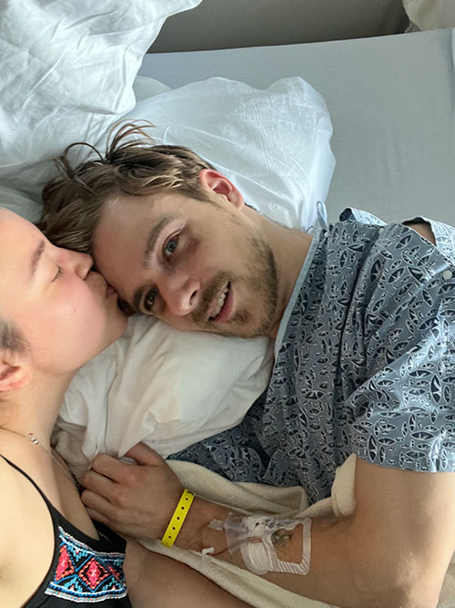 Noah smiles while his girlfriend kisses his forehead.