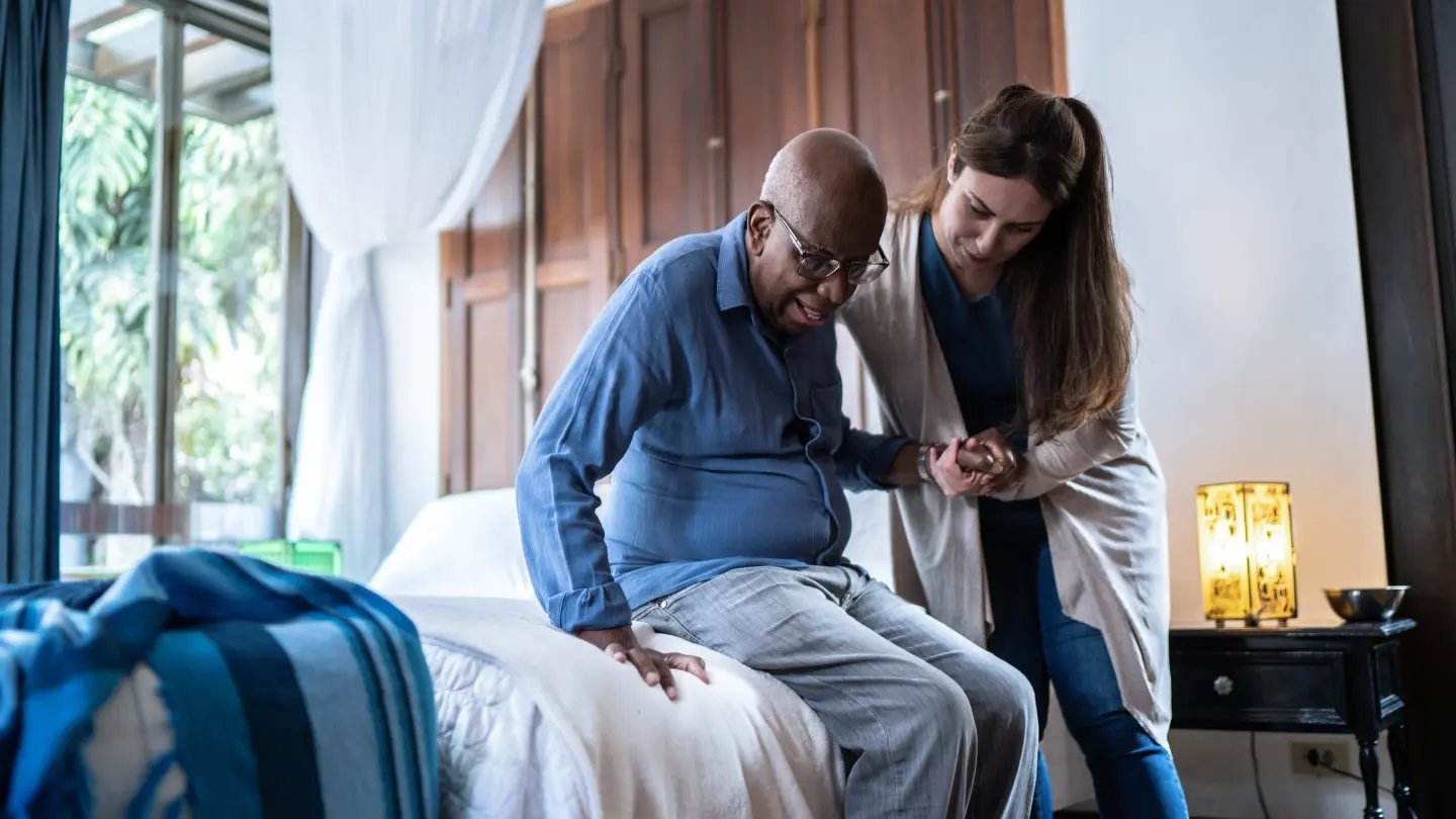 A female caregiver helping a senior man stand up from his bed at home.